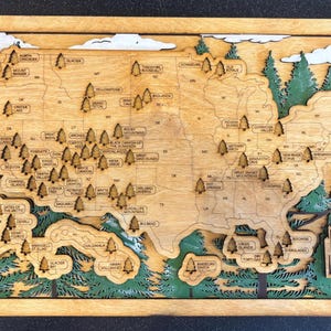 May include: A wooden map of the United States with a green and brown color scheme. The map features the names of all the national parks in the United States. The map is framed in a light brown wood frame. The text "Our National Park Adventure" is printed on the bottom right corner of the map.