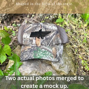 May include: Camouflage baseball cap with a black silhouette of a Bigfoot and a state outline. The cap has a mesh back and a curved brim. The text "Shaped in the form of the state" and "Two actual photos merged to create a mock up" are visible.