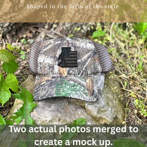 May include: Camouflage baseball cap with a Bigfoot silhouette and a flag design. The cap has a mesh back and a curved brim. The text "Shaped in the form of the state" and "Two actual photos merged to create a mock up" are also visible.