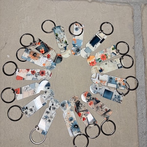 May include: A collection of 15 keychains with different colorful designs. The keychains are made of metal and have a ring attached to them. The designs feature flowers, abstract patterns, and vintage-inspired illustrations. The keychains are arranged in a circle on a light gray surface.