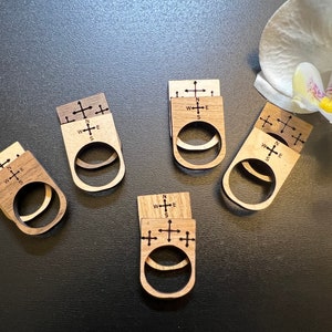 May include: Five wooden rings with a compass design. Each ring has a cross with the letters N, S, E, and W engraved on it. The rings are stacked on top of each other.