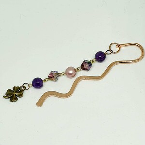 Könnte beinhalten: Ein Kupfer-Lesezeichen mit wellenförmigem Design, verziert mit violetten und rosafarbenen Perlen und einem Glückskleeblatt-Anhänger.