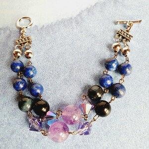 May include: A two-strand bracelet with silver-toned metal clasps and beads. The bracelet features blue lapis lazuli beads, black onyx beads, and amethyst beads with iridescent crystal accents.