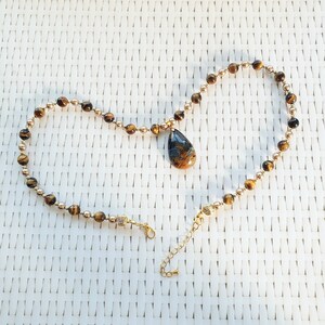May include: A gold necklace with a teardrop pendant. The necklace has small, round, gold beads and larger, brown tiger's eye beads. The pendant is a mix of brown and blue. The necklace has a clasp and an adjustable chain. The necklace is approximately 40 cm long.