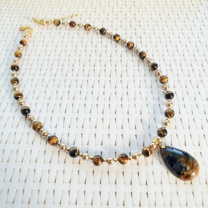 May include: A necklace with gold-coloured and tiger eye beads. The necklace has a teardrop-shaped pendant with a dark blue and brown pattern. The necklace is displayed on a white woven surface.