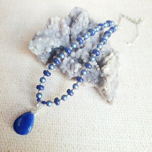 May include: A blue gemstone necklace with a teardrop pendant. The necklace is made of small, round beads and is strung on a silver chain.