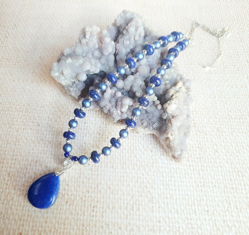 May include: A blue gemstone necklace with a teardrop pendant. The necklace is made of small, round beads and is strung on a silver chain.