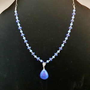 May include: A silver chain necklace with blue glass beads and a teardrop-shaped pendant. The pendant is a light blue color with a darker blue center.