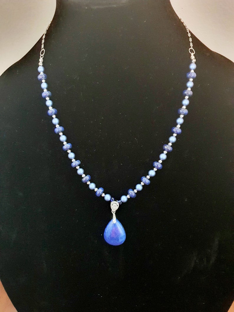 May include: A silver chain necklace with blue glass beads and a teardrop-shaped pendant. The pendant is a light blue color with a darker blue center.