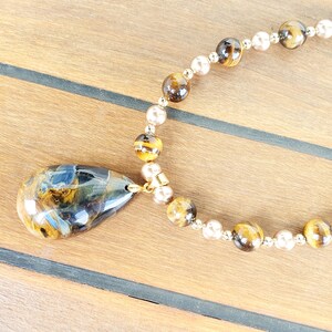May include: A necklace with a large, teardrop-shaped pendant in blue, brown, and gold tones. The necklace has alternating round beads in gold and pearl colours, connected by a gold chain.