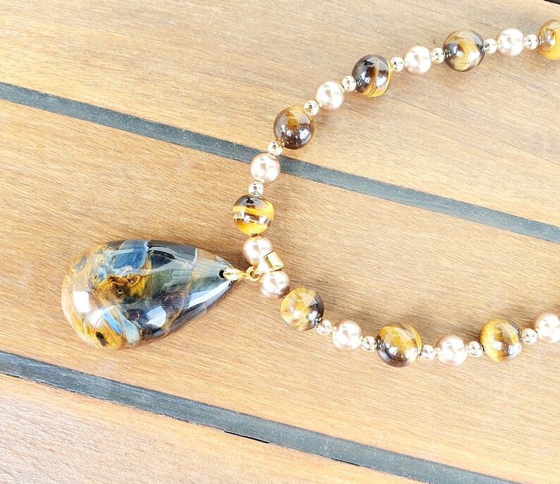 May include: A necklace with a large, teardrop-shaped pendant in blue, brown, and gold tones. The necklace has alternating round beads in gold and pearl colours, connected by a gold chain.