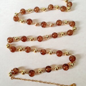 May include: A long gold chain necklace with brown glass beads and gold accents. The necklace is laid out in an S shape on a white surface.