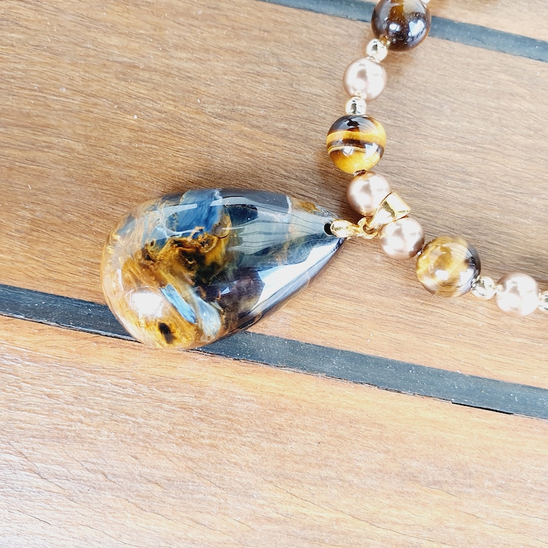 May include: A pendant necklace with a teardrop-shaped resin pendant. The pendant is a mix of blue and brown hues. The necklace features brown, gold, and pearl-coloured beads. The necklace is on a wooden surface.