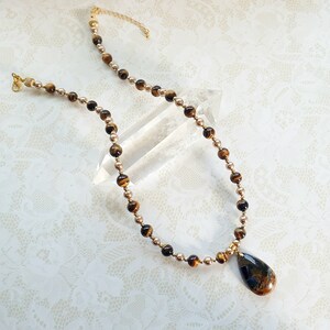 May include: A necklace with a teardrop pendant and a beaded chain. The pendant is a mix of brown and blue colours. The chain alternates between small gold-coloured beads and larger tiger's eye beads. The necklace has a gold-coloured clasp.