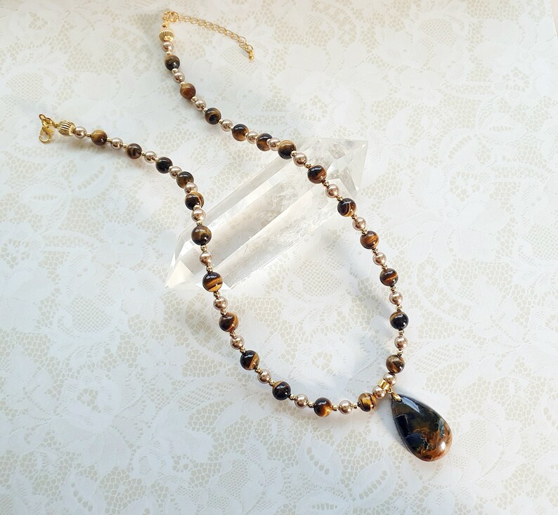 May include: A necklace with a teardrop pendant and a beaded chain. The pendant is a mix of brown and blue colours. The chain alternates between small gold-coloured beads and larger tiger's eye beads. The necklace has a gold-coloured clasp.