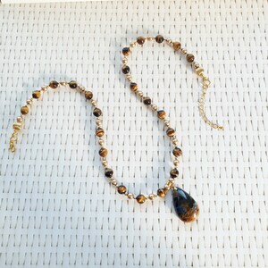 May include: A gold necklace with alternating tiger's eye and gold beads. A teardrop pendant with a dark blue and brown pattern is suspended from the centre. The necklace is displayed on a white woven surface.