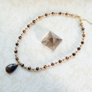 May include: A necklace with tiger's eye and gold-coloured beads. A teardrop-shaped pendant is attached to the necklace. A clear, pyramid-shaped crystal is placed nearby. The necklace is displayed on a white, lace-textured surface.