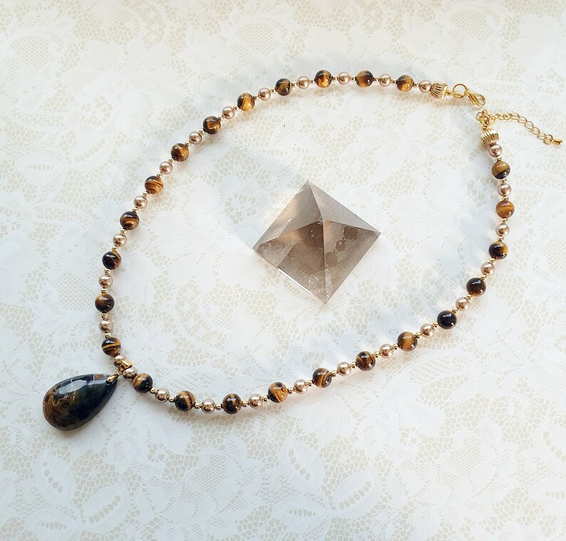 May include: A necklace with tiger's eye and gold-coloured beads. A teardrop-shaped pendant is attached to the necklace. A clear, pyramid-shaped crystal is placed nearby. The necklace is displayed on a white, lace-textured surface.