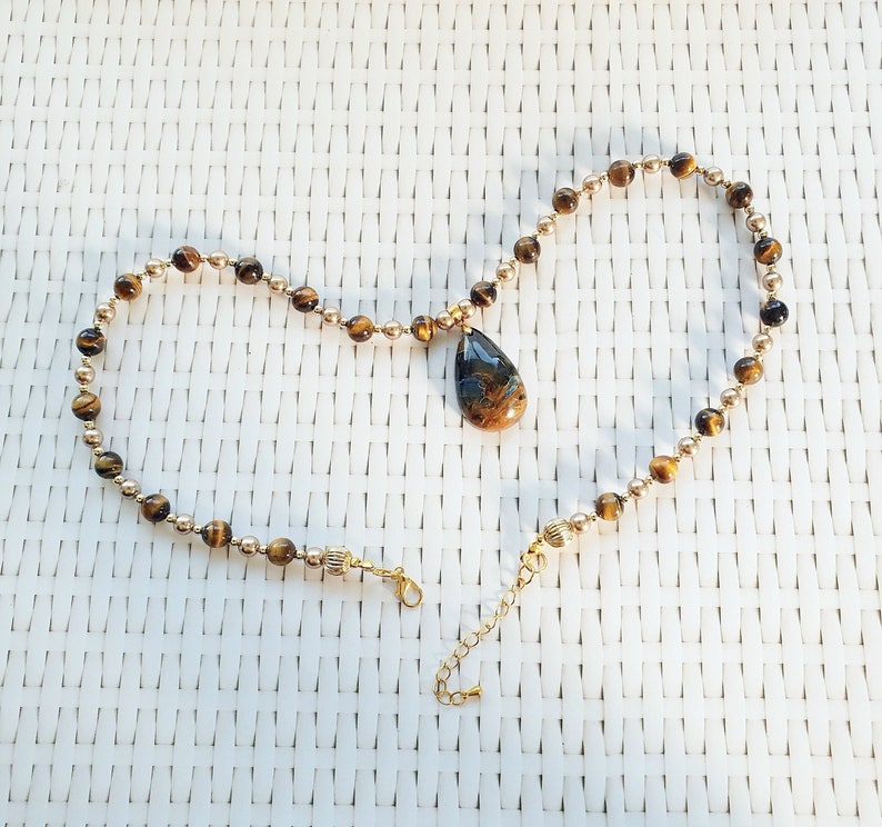 May include: A gold necklace with a teardrop pendant. The necklace has small, round, gold beads and larger, brown tiger's eye beads. The pendant is a mix of brown and blue. The necklace has a clasp and an adjustable chain. The necklace is approximately 40 cm long.