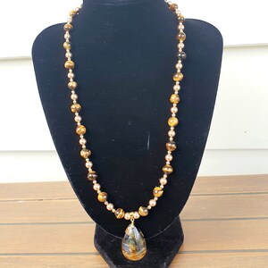 May include: A long necklace with alternating tiger's eye and gold-coloured beads. A teardrop-shaped pendant with a marbled pattern hangs from the centre. The necklace is displayed on a black velvet bust.