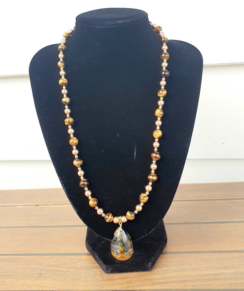 May include: A long necklace with alternating tiger's eye and gold-coloured beads. A teardrop-shaped pendant with a marbled pattern hangs from the centre. The necklace is displayed on a black velvet bust.