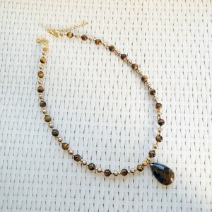 May include: A gold necklace with alternating small gold beads and larger, round, brown tiger eye beads. A teardrop-shaped tiger eye pendant hangs from the centre. The necklace is displayed on a white woven surface.