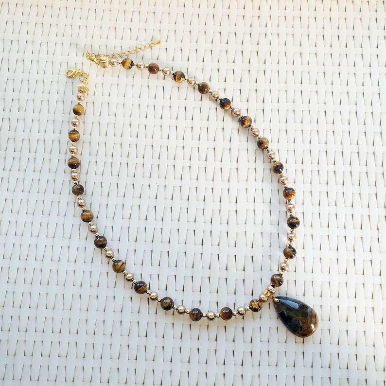 May include: A gold necklace with alternating small gold beads and larger, round, brown tiger eye beads. A teardrop-shaped tiger eye pendant hangs from the centre. The necklace is displayed on a white woven surface.