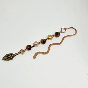 May include: A copper bookmark with a wavy design and a chain of tiger's eye and pearl beads. A small leaf charm hangs from the end of the chain.
