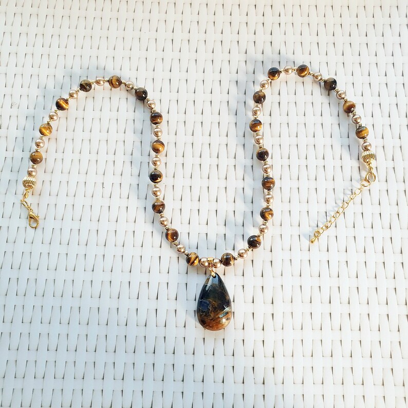 May include: A necklace with a teardrop pendant and a beaded chain. The gold-coloured chain features alternating brown and gold beads. The pendant is a dark blue and brown colour.
