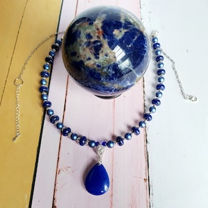 May include: A blue gemstone necklace with a teardrop pendant. The necklace is made of small, round beads and has a silver chain. The pendant is a large, teardrop-shaped gemstone with a silver setting.
