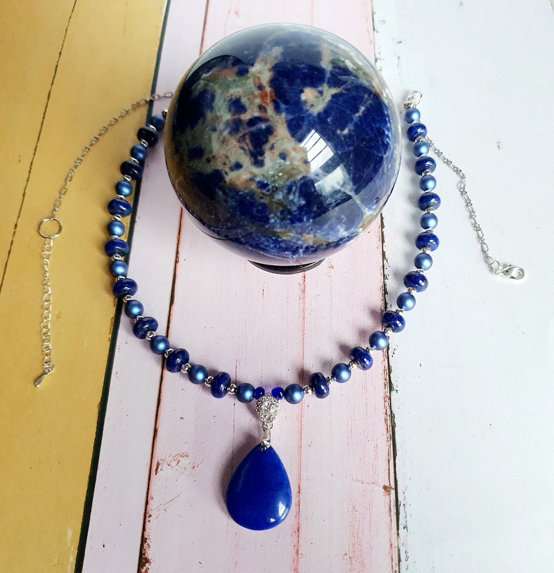 May include: A blue gemstone necklace with a teardrop pendant. The necklace is made of small, round beads and has a silver chain. The pendant is a large, teardrop-shaped gemstone with a silver setting.
