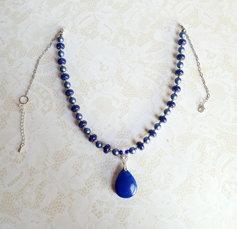 May include: A silver chain necklace with a teardrop-shaped lapis lazuli pendant. The necklace is made with blue and light blue glass beads.