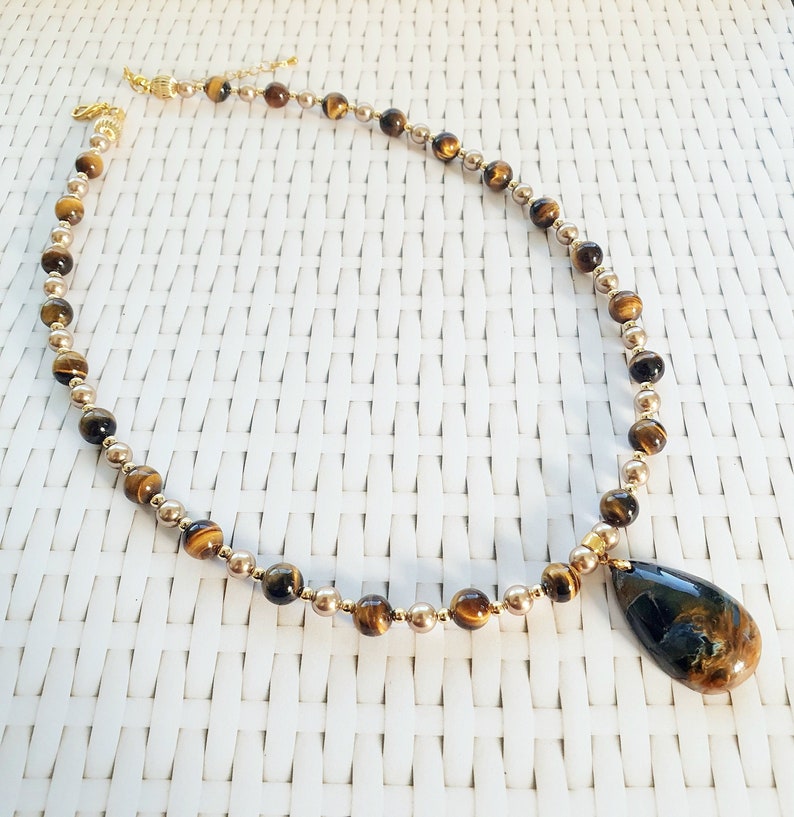 May include: A necklace with gold-coloured and tiger eye beads. The necklace has a teardrop-shaped pendant with a dark blue and brown pattern. The necklace is displayed on a white woven surface.