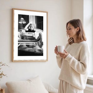 May include: A framed monochrome print featuring a woman reading a newspaper with the word "DAILY" visible. The print is in a light wood frame. A woman in a cream sweater holds a white mug, standing beside the framed print.