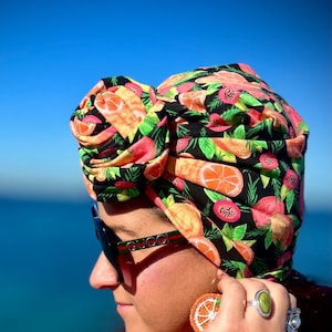 Op de afbeelding: Een vrouw draagt een kleurrijke turban met een citrusfruitprint, een zonnebril en een turquoise kralenarmband. Ze draagt een rood topje en een citrusfruit-oorring.