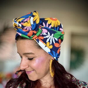 May include: A woman with long dark hair is wearing a colourful floral print turban. The turban is tied in a knot on top of her head. She is also wearing large yellow hoop earrings.