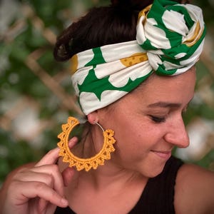 May include: A woman wearing a white and green patterned headscarf and a black top. She is wearing large, yellow crochet hoop earrings. The earrings have a sunburst design.