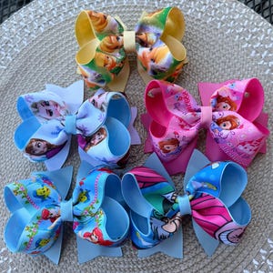 May include: A collection of six colorful hair bows. The bows feature various cartoon characters, including princesses and other animated figures. The bows have a layered design with a mix of light blue, pink, yellow, and other vibrant colors.