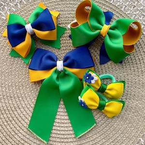Può includere: Una collezione di cinque fiocchi per capelli in una palette di colori verde, giallo e blu navy. I fiocchi sono realizzati in tessuto a coste, alcuni con code lunghe, nodi centrali bianchi e piccoli accenti floreali decorativi con perline.