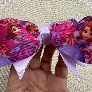 May include: A hair bow made of ribbed fabric featuring a repeating print of a cartoon princess in a purple dress with a tiara, surrounded by pink roses. The bow is secured in the center with a light purple ribbon and has pointed ends.