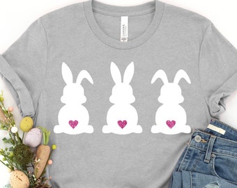 May include: A gray t-shirt with three white bunny silhouettes, each with a pink heart on its backside. The shirt is laid out on a white background with a pair of white sneakers, blue jeans, a carrot, and a nest with Easter eggs.