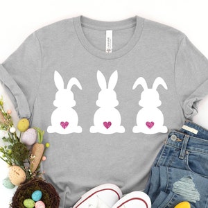 May include: A gray t-shirt with three white bunny silhouettes, each with a pink heart on its backside. The shirt is laid out on a white background with a pair of white sneakers, blue jeans, a carrot, and a nest with Easter eggs.