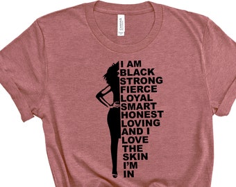 May include: A dusty rose t-shirt with a black silhouette of a woman and the words "I AM BLACK STRONG FIERCE LOYAL SMART HONEST LOVING AND I LOVE THE SKIN I'M IN" printed vertically on the right side.