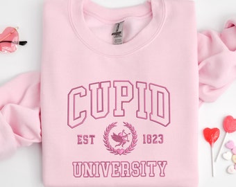 May include: Pink sweatshirt with the text "CUPID UNIVERSITY" in a vintage collegiate style font. The text is in a darker pink color and includes the words "EST 1823" and a graphic of a winged cupid with an arrow.