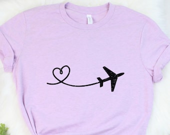 Puede incluir: Una camiseta lavanda con un avión negro y un gráfico de corazón. El avión está volando lejos de un corazón.