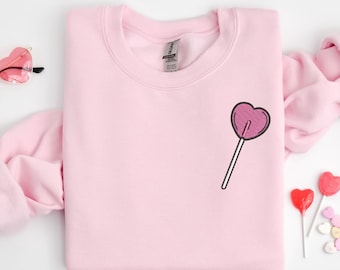 May include: A pink sweatshirt with a heart-shaped lollipop graphic on the front. The sweatshirt is folded and laid flat on a white surface.