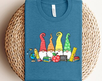 May include: Teal t-shirt with a graphic of five gnomes wearing colourful hats, holding school supplies. The gnomes are in front of books, an apple, and a chalkboard with "ABC" on it. A pencil, globe, and ruler are also included.