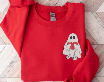 Puede incluir: Una sudadera roja con un fantasma blanco sosteniendo un corazón rojo. El fantasma tiene ojos negros y está bordado en la parte delantera. La sudadera tiene cuello redondo y mangas largas.