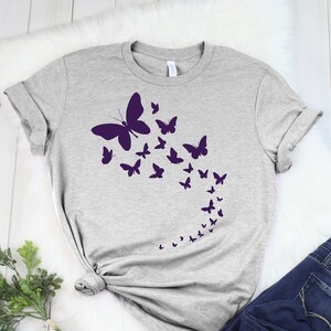 May include: A light gray t-shirt featuring a design of purple butterflies in various sizes, arranged in a flowing pattern. The butterflies appear to be in flight. The shirt is paired with dark blue jeans.