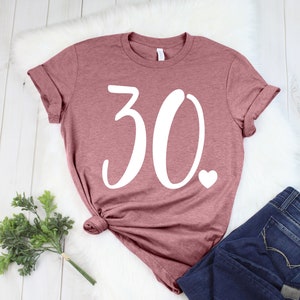 May include: A mauve t-shirt featuring the number "30" in white script and a small white heart. The shirt is styled with dark blue jeans. The shirt is knotted at the bottom.